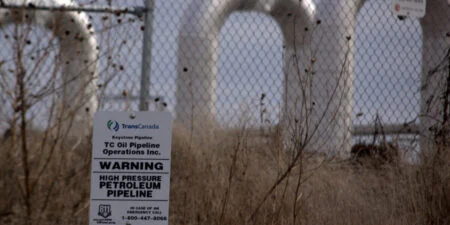 Canada’s Biggest Pension Fund Expands Fossil Fuel Investments Despite Climate Warnings Warning sign reading “High Pressure Petroleum Pipeline” on a chain-link fence with large pipeline infrastructure visible in the background amid dry grass and weeds.