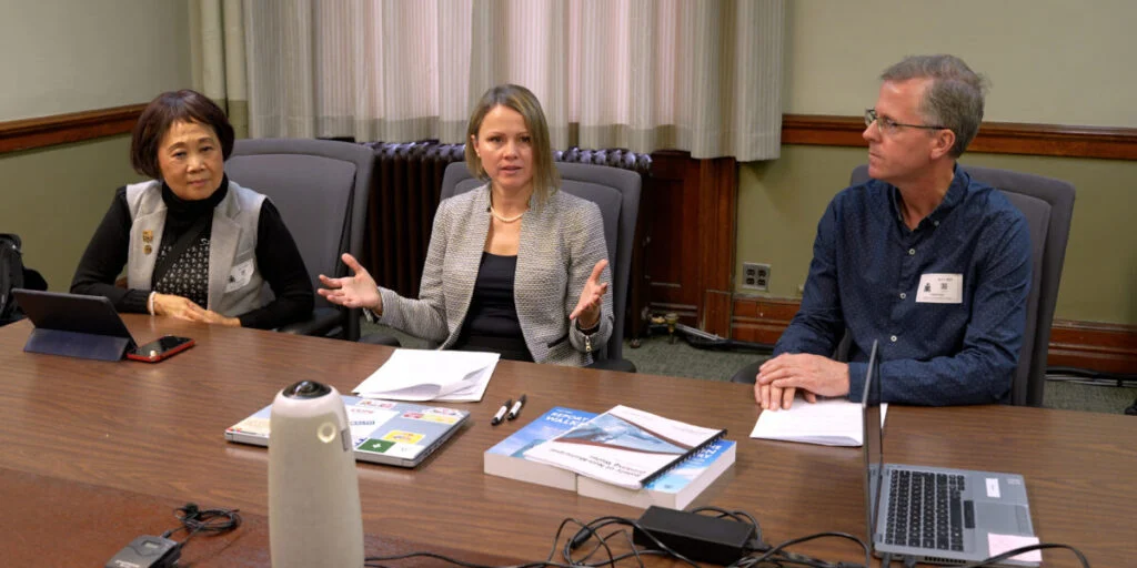 Three people sit at a conference table during a meeting. The woman in the center speaks while gesturing with her hands, with documents, laptops, and devices on the table in front of them. The two other participants listen on either side in a boardroom setting.