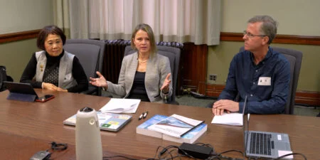 Ontario Public Health Funding 2026 — Crisis Warnings Grow As Cuts Bite Three people sit at a conference table during a meeting. The woman in the center speaks while gesturing with her hands, with documents, laptops, and devices on the table in front of them. The two other participants listen on either side in a boardroom setting.