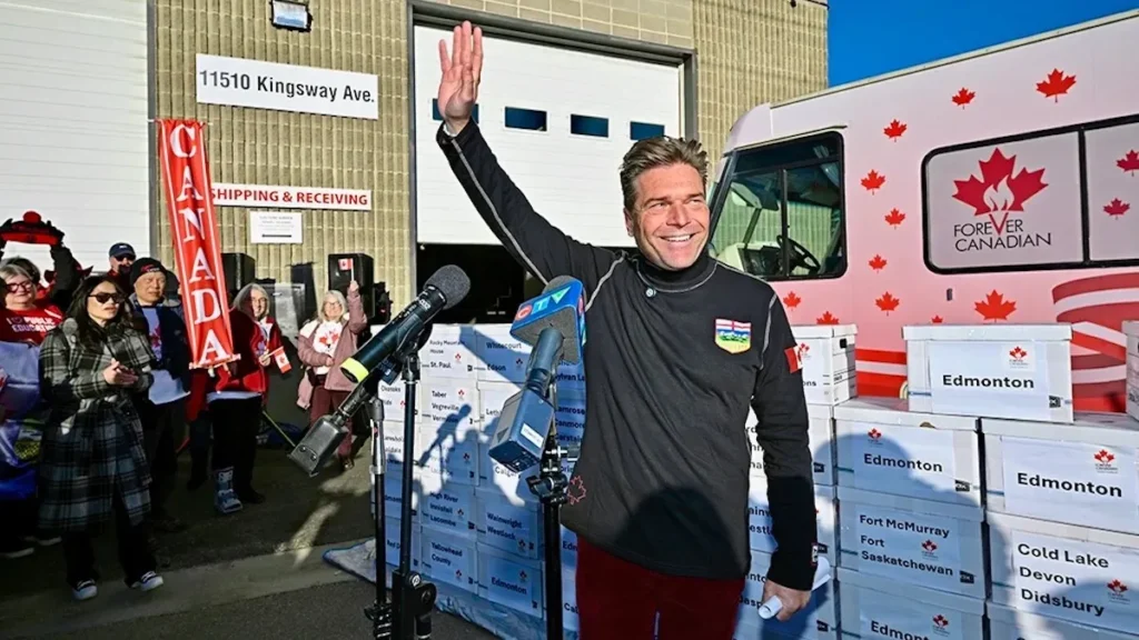 Alberta Unity Petition Approved — Elections Alberta Clears The Way For A Province-Wide Signature Drive A smiling man stands outdoors waving beside microphones during a public event, with a group of supporters behind him holding Canadian flags and banners. He is positioned in front of stacked boxes labeled with Alberta communities, and a vehicle decorated with red maple leaves and the “Forever Canadian” logo is visible on the right. The scene appears to be part of a campaign or petition delivery related to Alberta’s place in Canada.