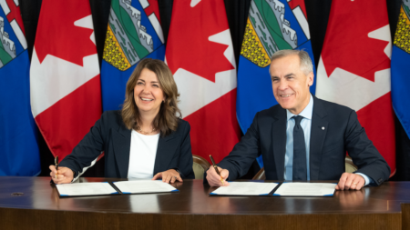 Alberta Poised For Early Election As Prince Rupert Pipeline Falters A woman and a man sit at a table, smiling as they sign documents in front of a backdrop of Canadian and Alberta flags.