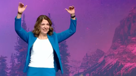 Danielle Smith Booed At Ucp Convention — Signs Of Deep Division Within Party A woman in a blue suit raises both hands while standing on a stage with a purple mountain landscape backdrop.