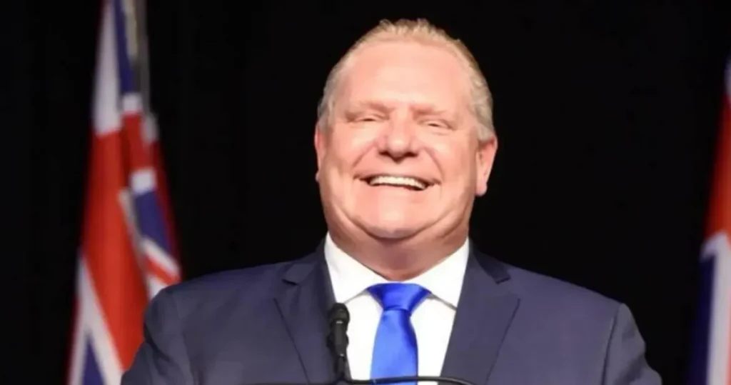 Ontario Water Under Threat: Critics Say Ford’s Policies Push Province Toward Privatization A smiling middle-aged man in a dark suit and bright blue tie stands at a podium, with two Canadian provincial-style flags blurred in the background against a dark backdrop.