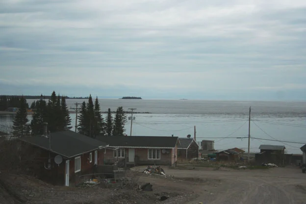 $908 Cab Fare: The Harsh Reality Of Food Access In Northern Communities A small northern coastal community with a few modest houses in the foreground, surrounded by trees and dirt roads, overlooking a vast, calm body of water under a cloudy sky with distant islands visible on the horizon.